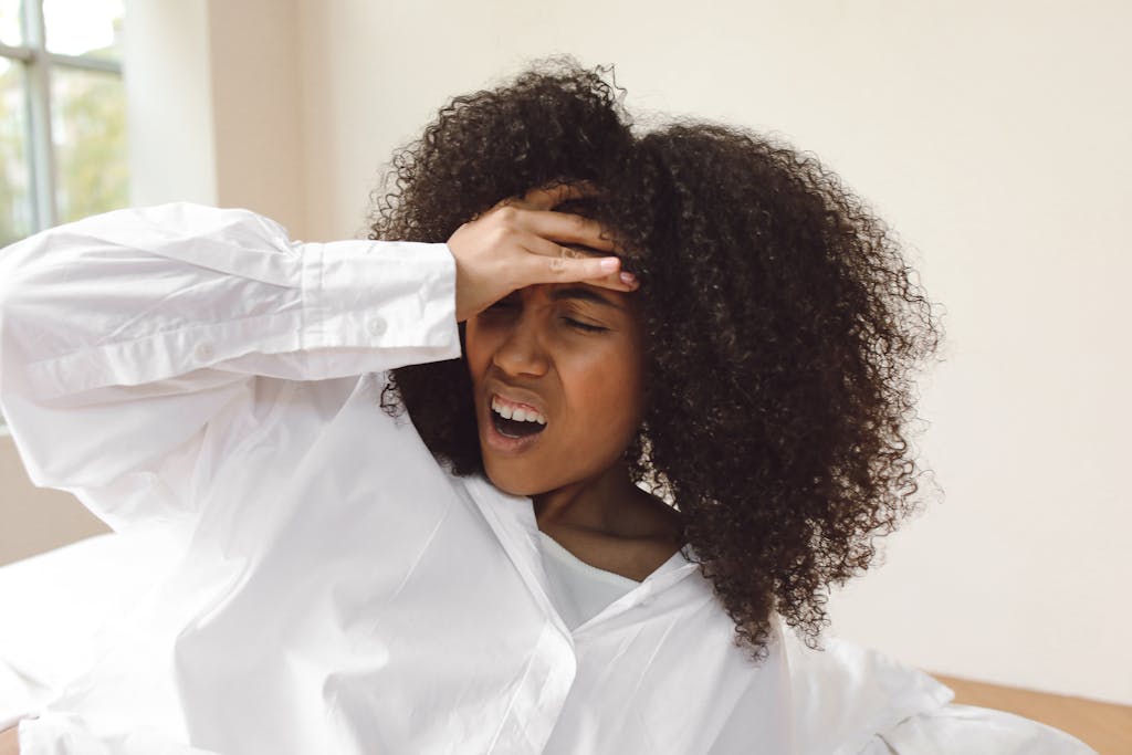 A young woman with curly hair waking up with a headache. She wears a white shirt and seems to be experiencing discomfort.